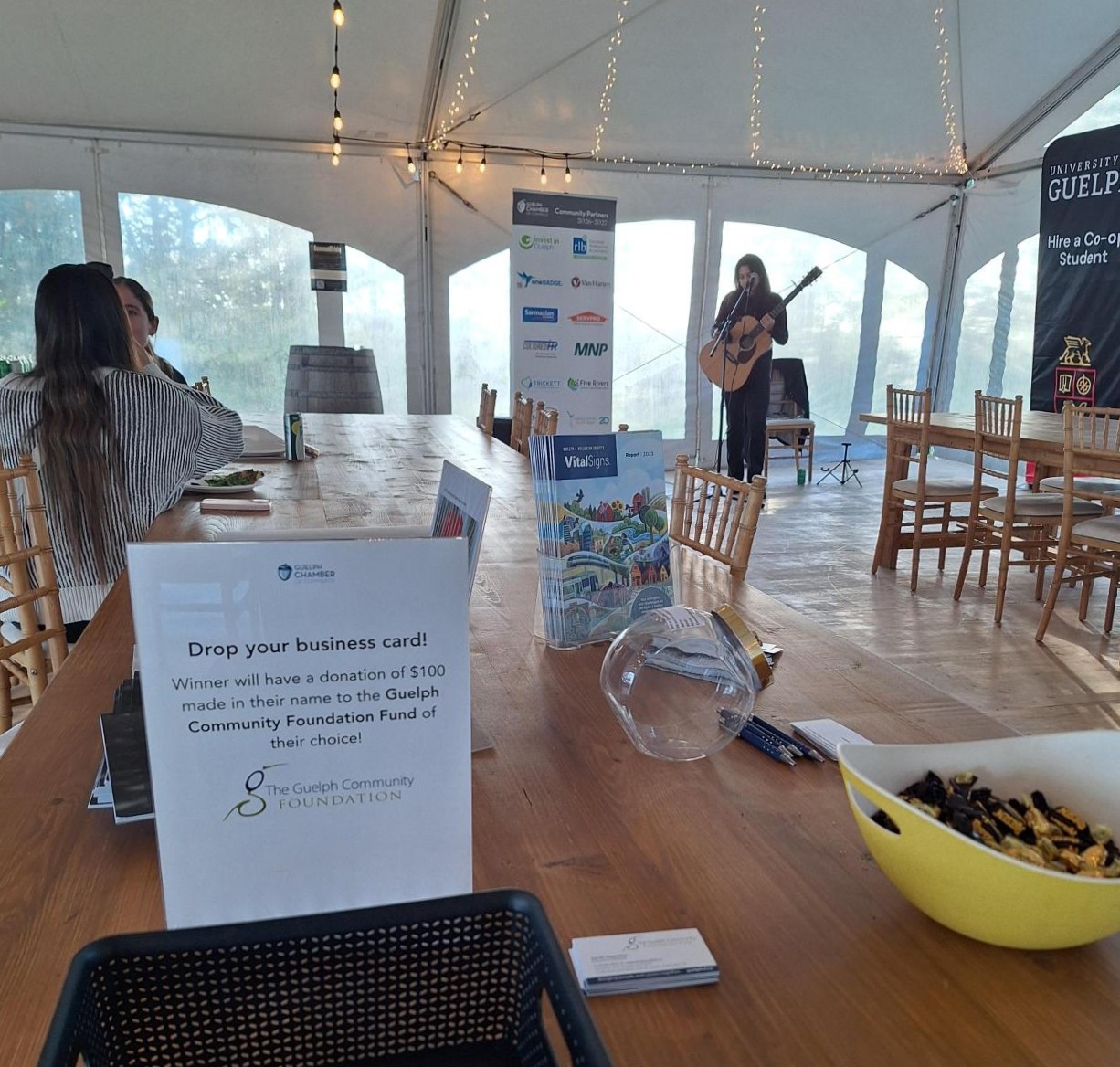 Indoor event space with a musician playing guitar in the background and a table featuring a business card drop box and snacks.
