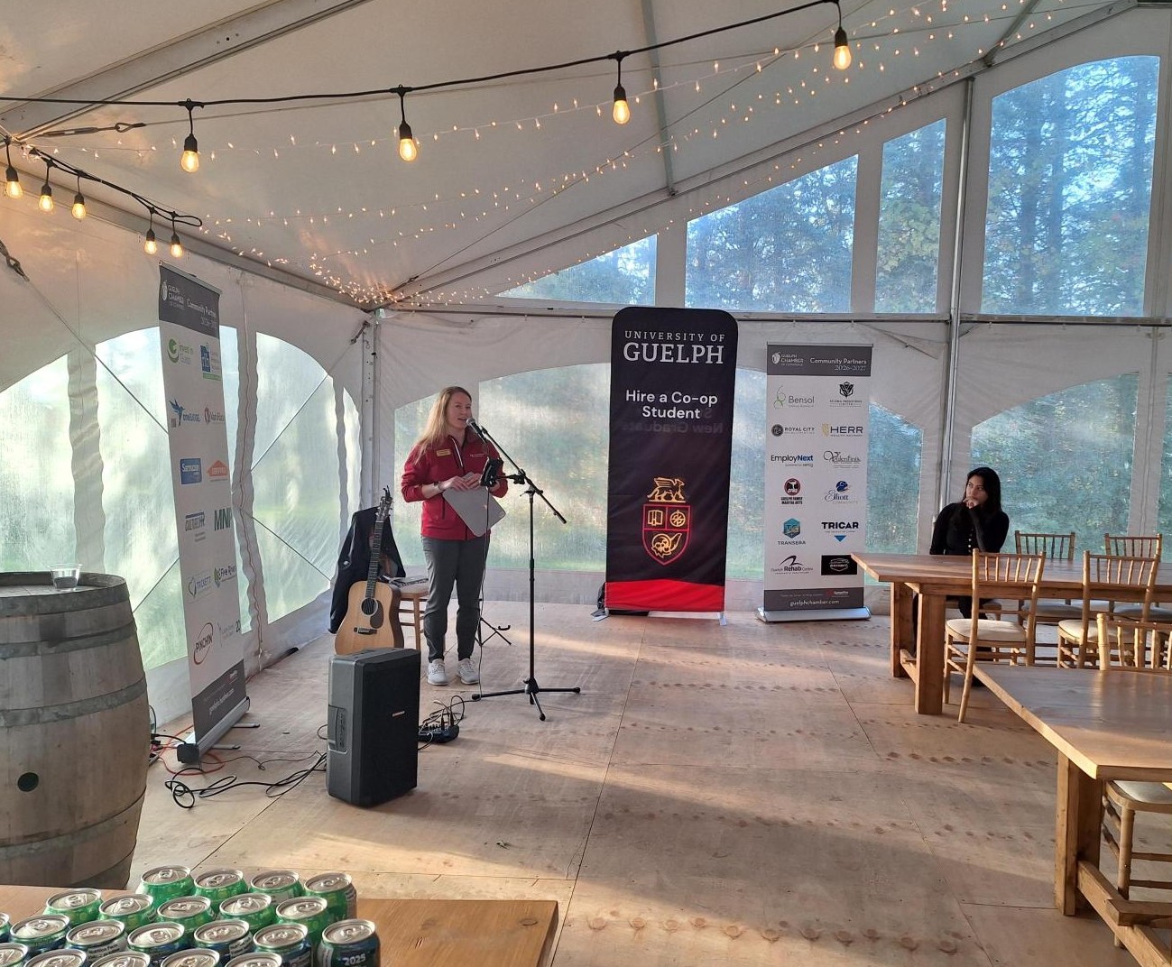 Woman speaking into a microphone inside a tent decorated with string lights and banners.