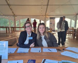 Two women sitting behind a registration table with sign-up sheets and informational brochures inside a large tent with string lights.