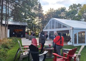 People socializing near a white event tent and wooden cabin surrounded by trees at dusk.