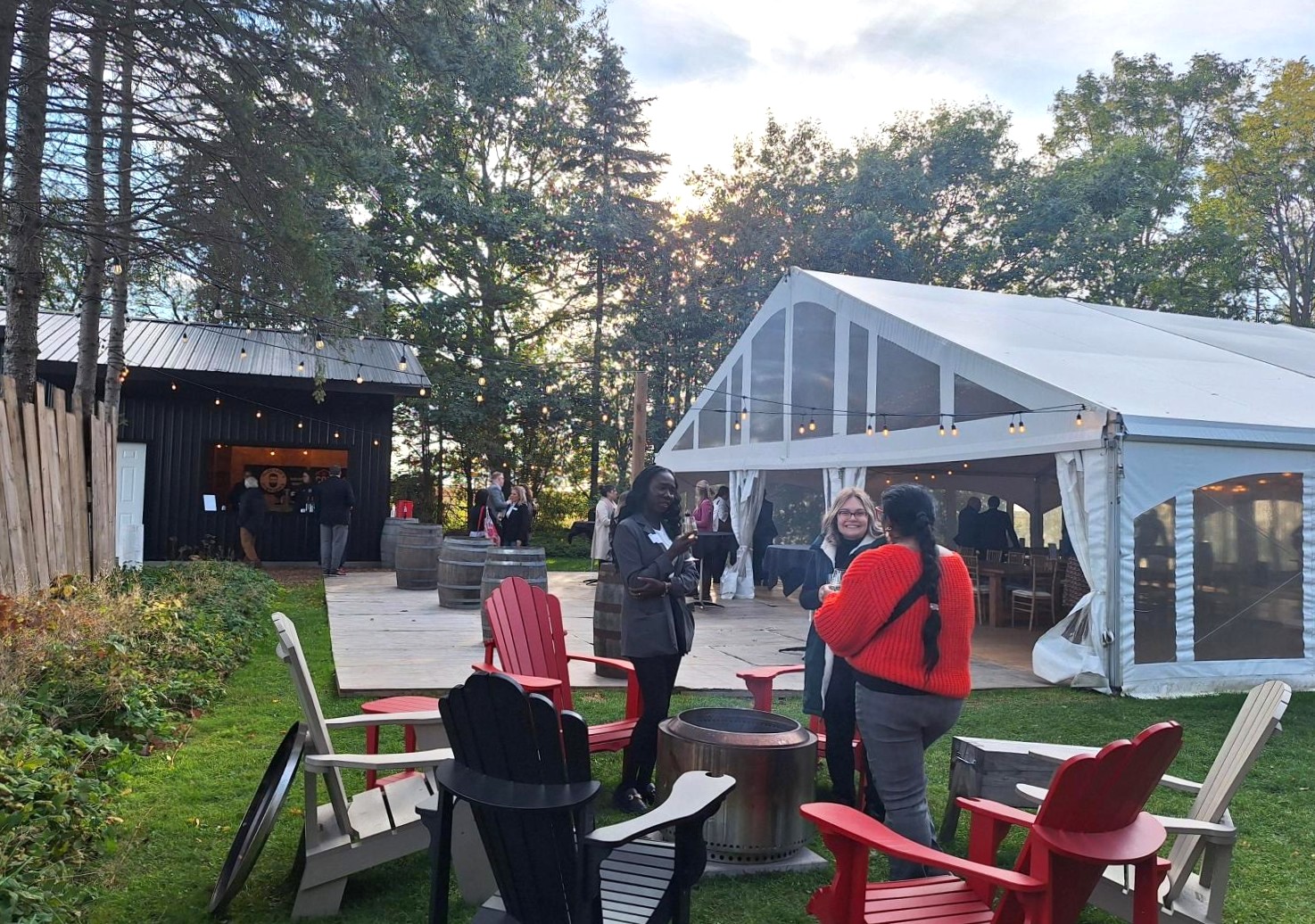 People socializing near a white event tent and wooden cabin surrounded by trees at dusk.
