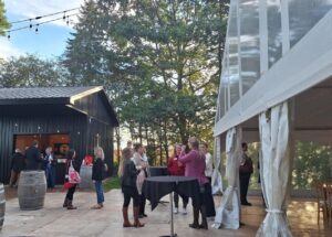 Guests socialize outdoors near a black barn-style building and a clear tent with white curtains during a sunny event.