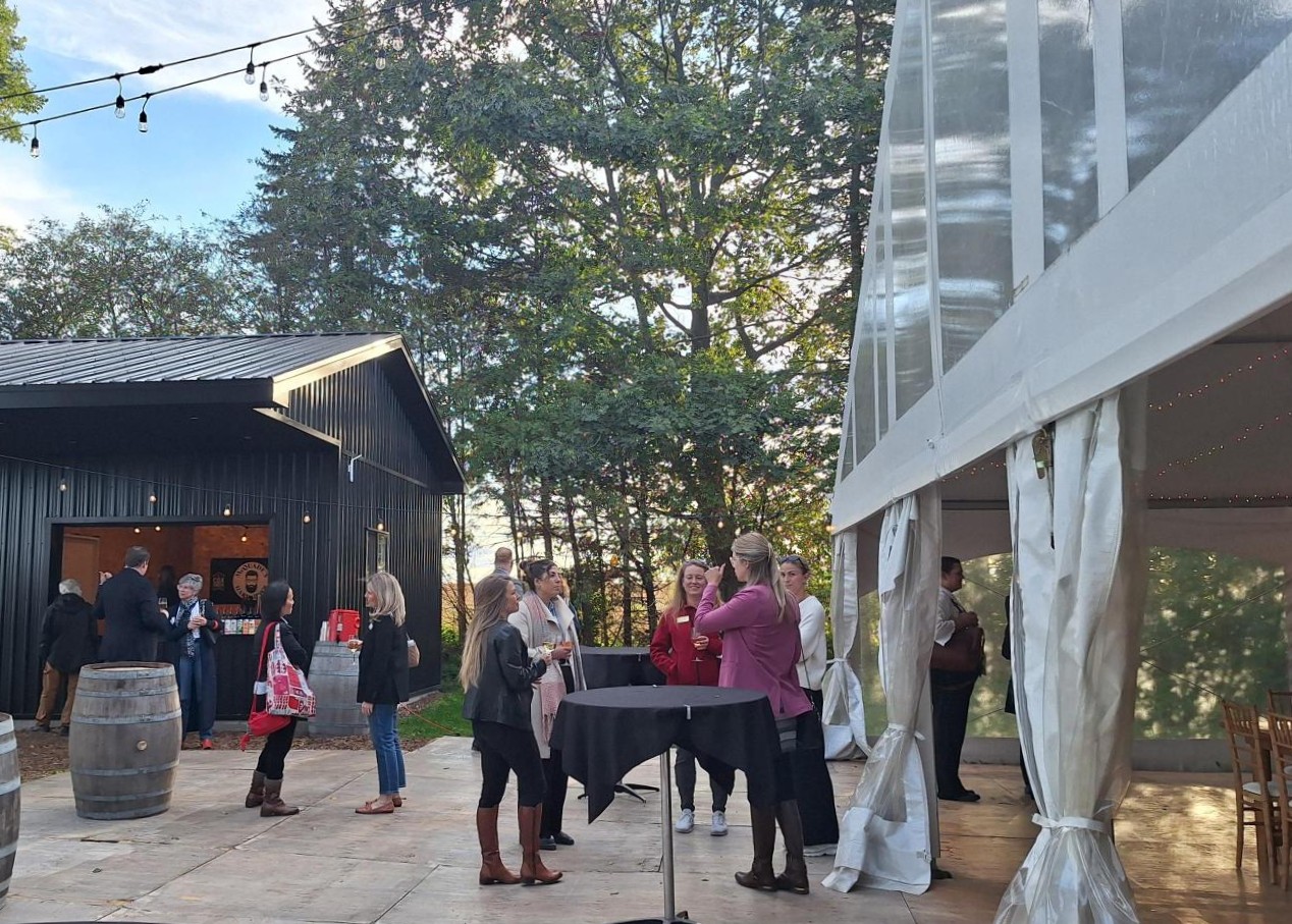 Guests socialize outdoors near a black barn-style building and a clear tent with white curtains during a sunny event.