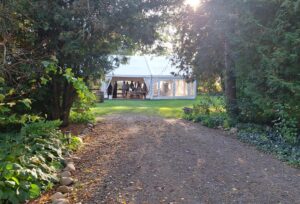 Gravel path leading through lush greenery to a white event tent with people gathered inside on a sunny day.