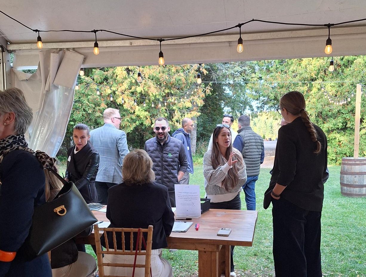 People socializing under a tent with string lights, surrounded by greenery and wooden barrels outdoors.