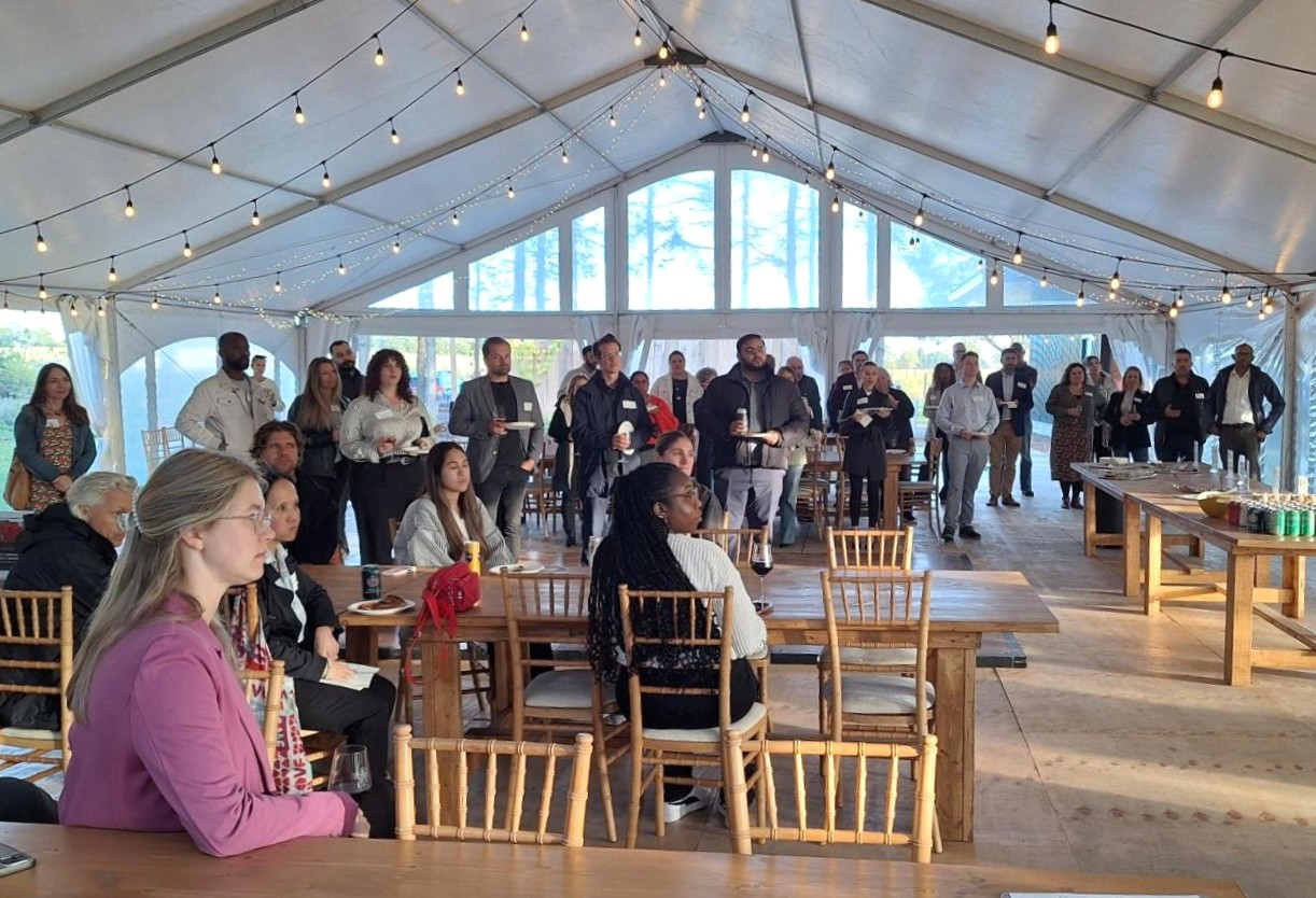 People gathered inside a large white tent with string lights, seated and standing around wooden tables during an event.