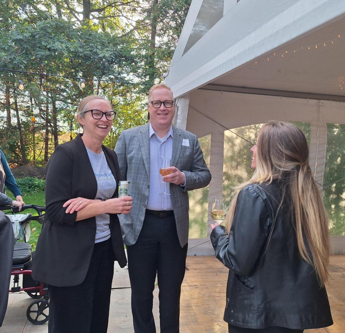 Three professionally dressed people smiling and holding drinks near a white event tent outdoors.
