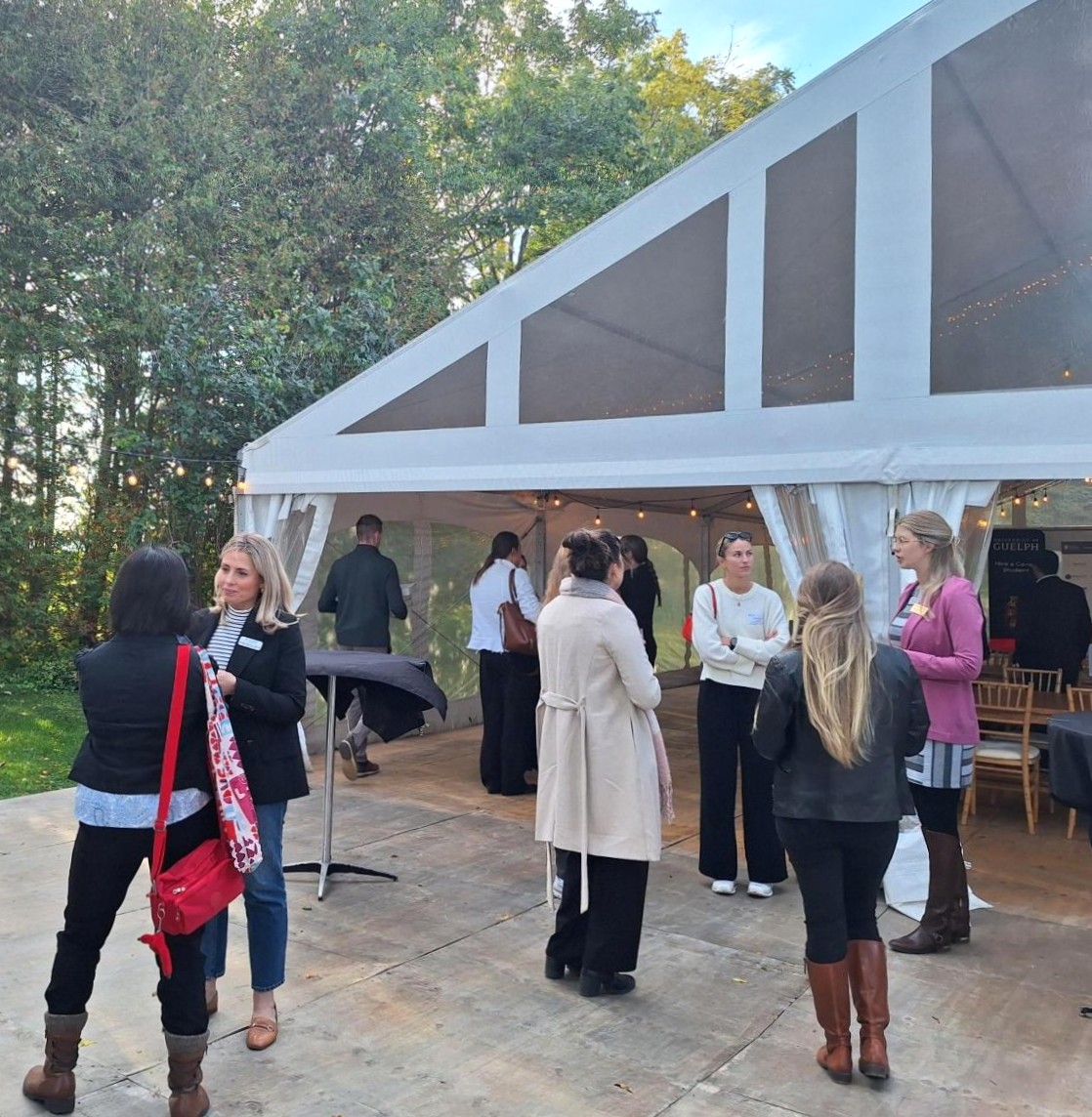 People socializing outside a white event tent surrounded by trees during a sunny day.