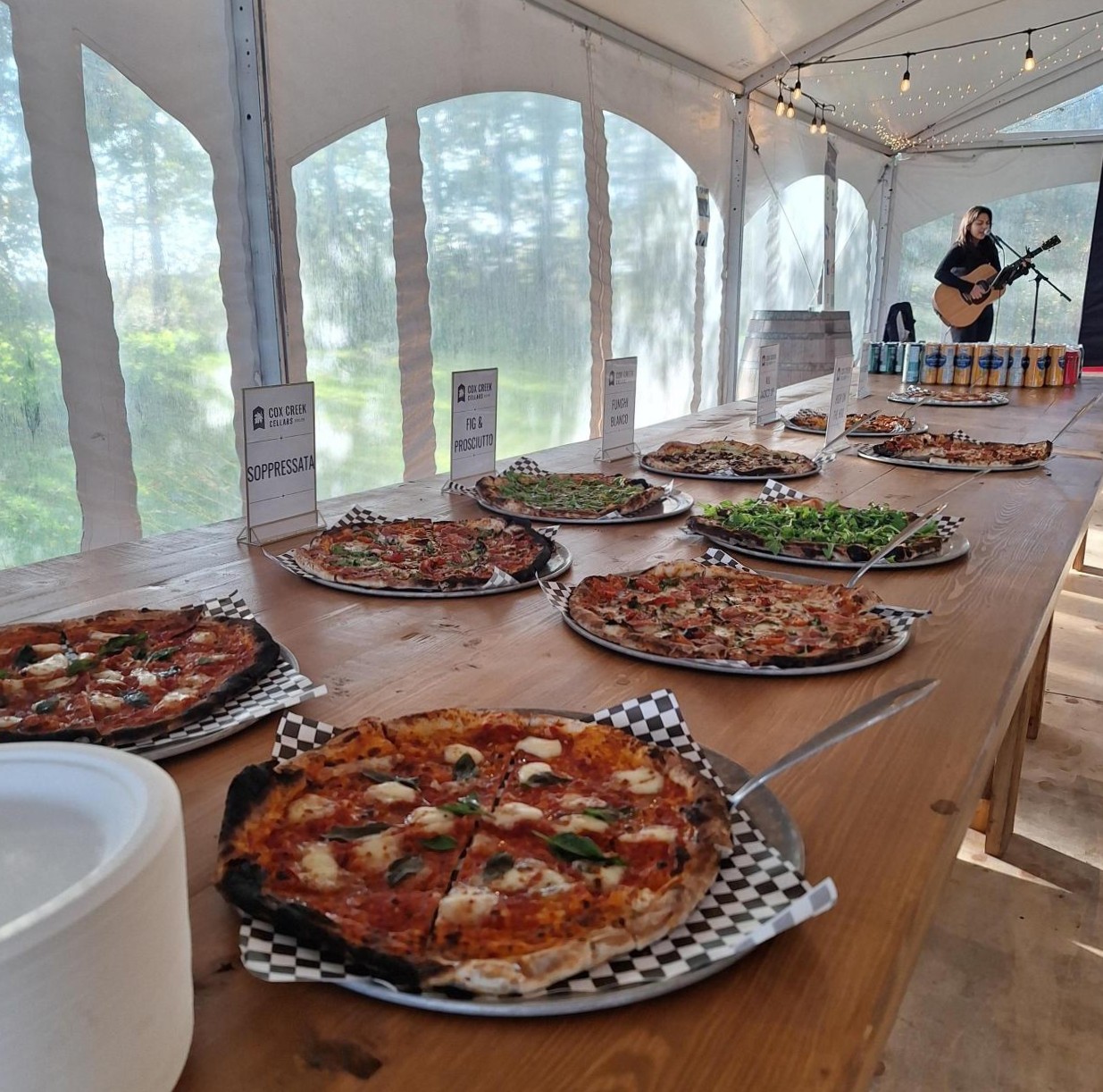Variety of freshly baked pizzas displayed on a wooden table inside a tent with string lights overhead and a musician playing guitar in the background.