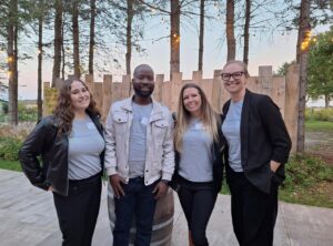 Four colleagues smiling outdoors in casual attire with trees and string lights in the background at dusk.