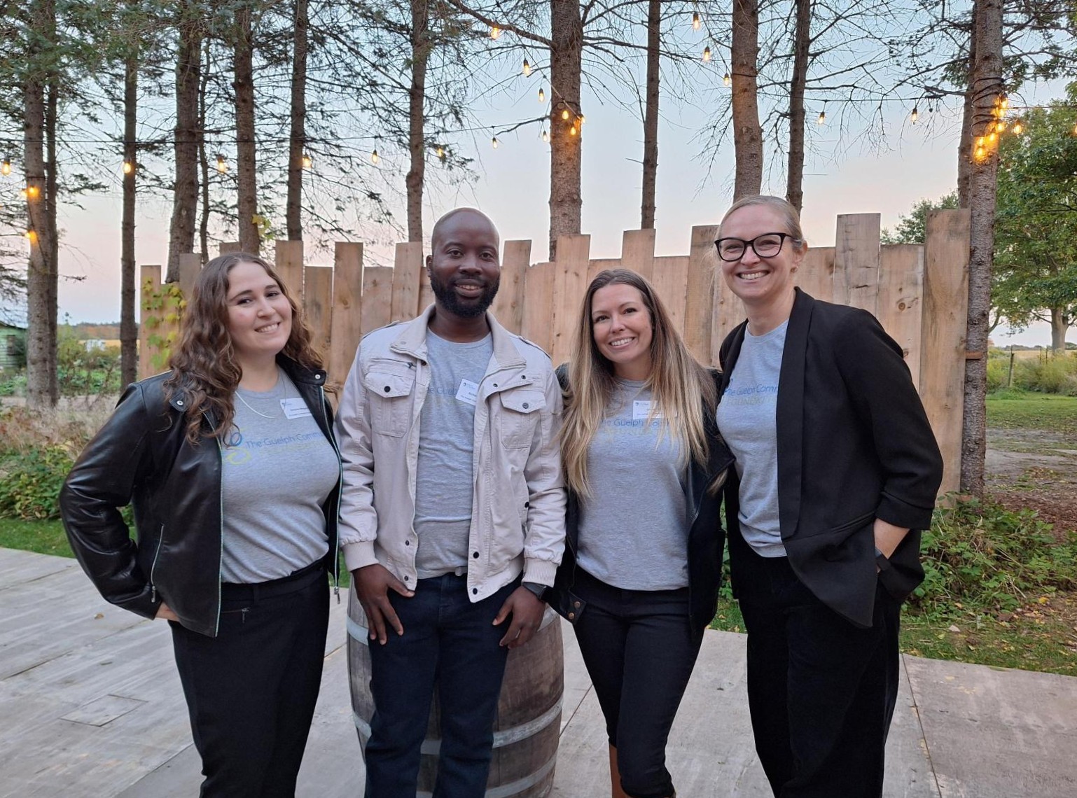 Four colleagues smiling outdoors in casual attire with trees and string lights in the background at dusk.